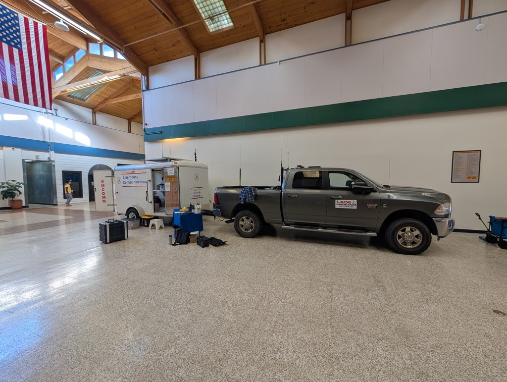 An indoor space featuring a large American flag, a parked gray pickup truck, and a trailer marked 'Emergency Communications.' Various equipment is set up beside the truck on a tiled floor.