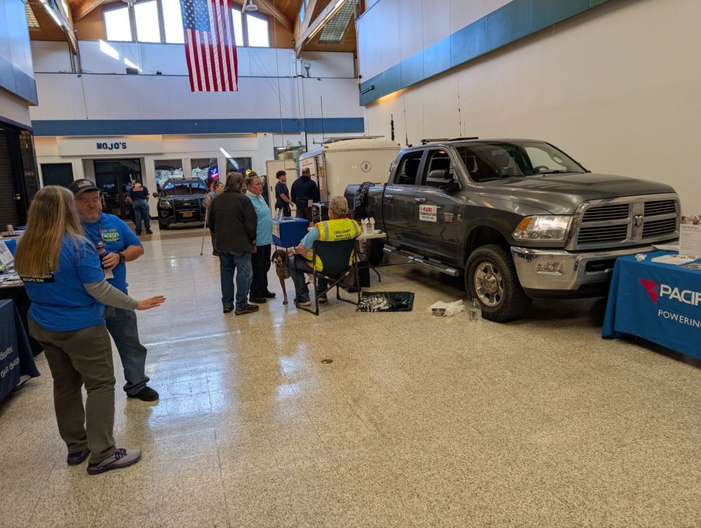 An indoor event with a gray pickup truck displayed alongside various booths. People are interacting, and an American flag is visible in the background.