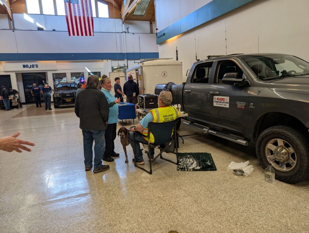 A gathering at a community event featuring a grey truck with communication equipment, attendees chatting, and a seated individual with a dog.