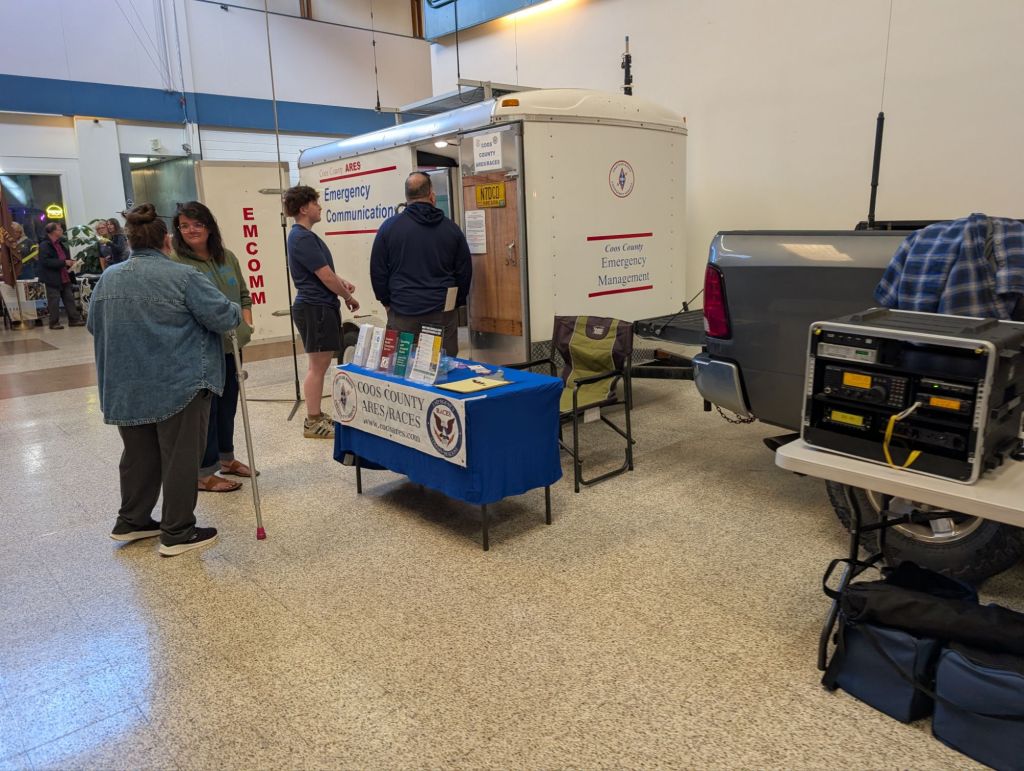 A group of people interacting at an emergency communication booth set up by Coos County ARES/RACES, featuring a trailer and equipment for emergency management.