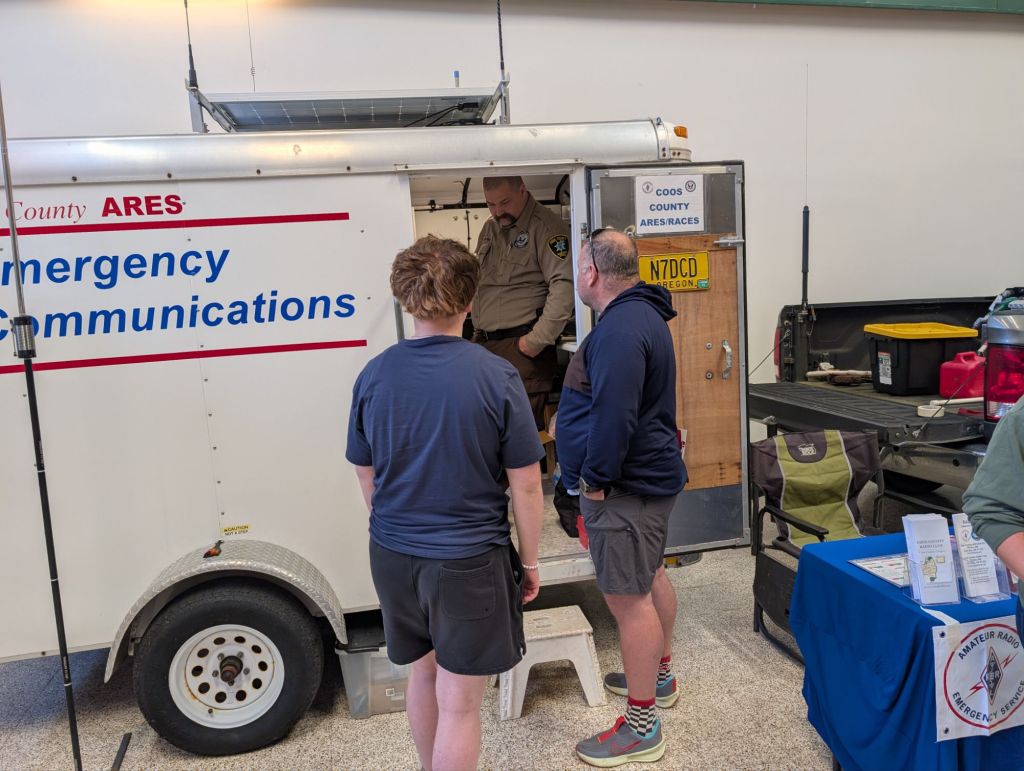 A group of people interacting with a mobile emergency communications trailer, with a uniformed officer inside. The scene includes informational materials on a table and a parked vehicle in the background.