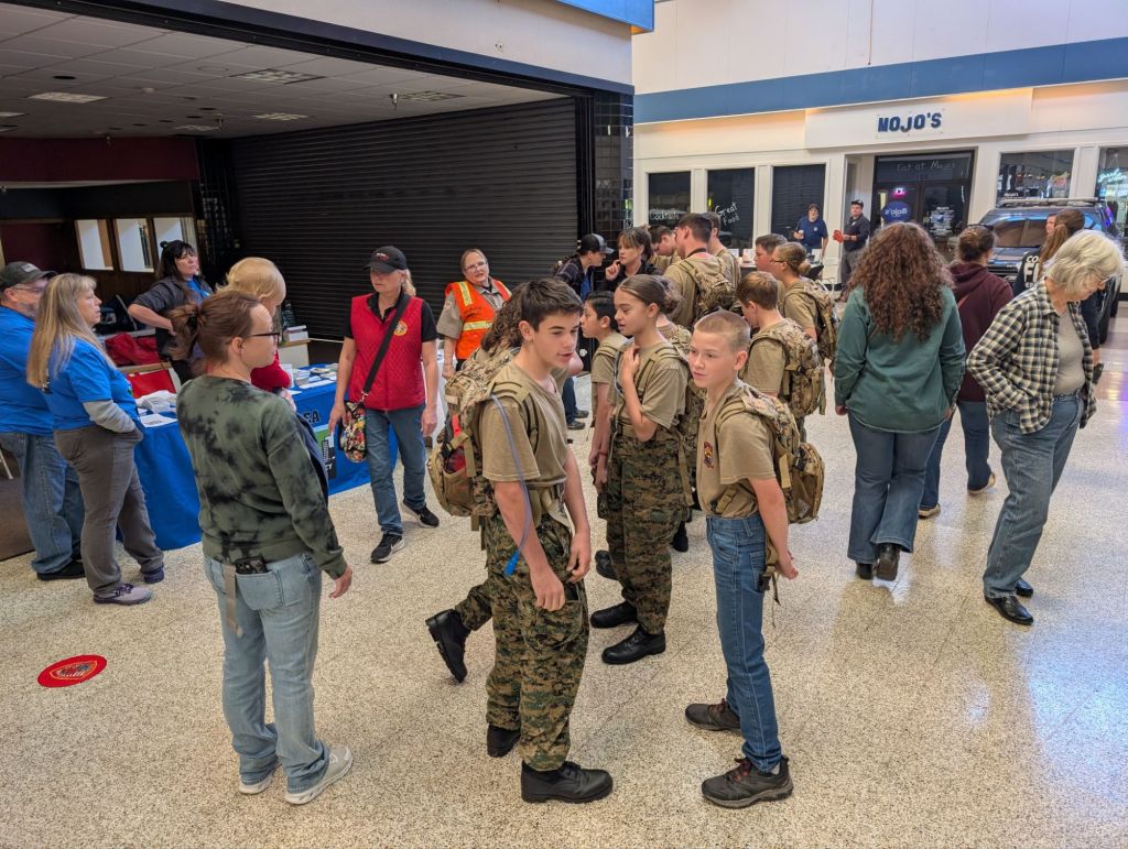 A group of young people in military-style outfits with backpacks gathers indoors, surrounded by adults wearing casual clothing. There are tables with informational materials in the background, and a variety of people engaged in conversation.