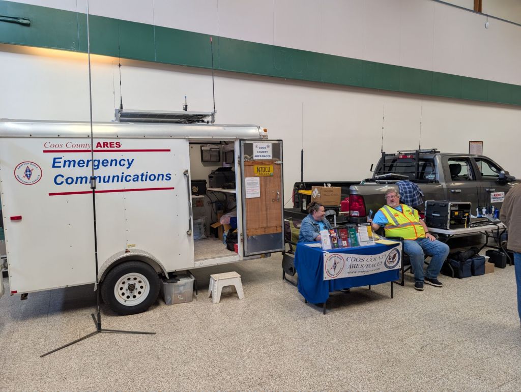 An emergency communications trailer with 'Coos County ARES' branding, accompanied by a table displaying informational materials and two people sitting behind it. A pickup truck is parked nearby.